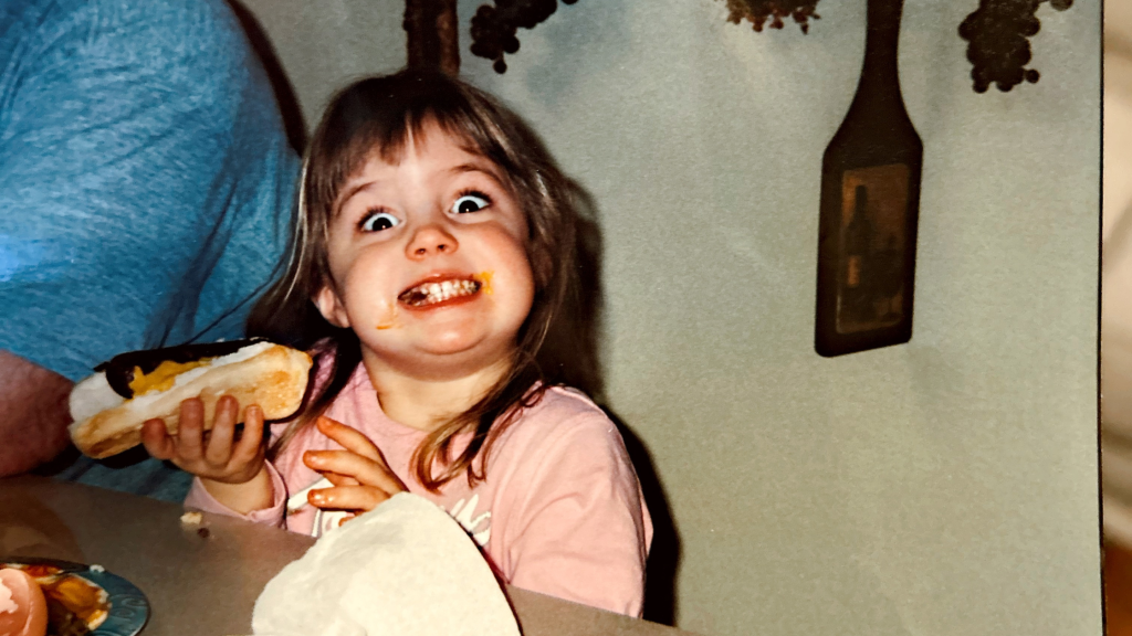 Emily as a toddler eating a hot dog and smiling