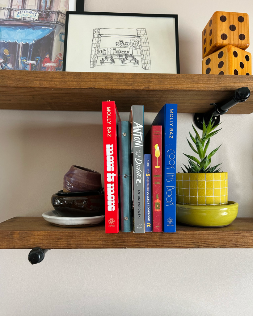 Wooden shelves with art, vintage dice, pottery pieces, a plant, and cookbooks. 